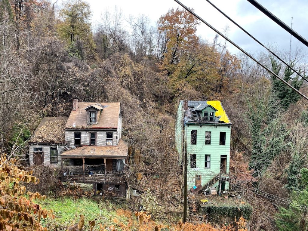 Two abandoned hillside houses beside the Rising Main public stairs in Pittsburgh, Pennsylvania, surrounded by overgrowth and autumn trees.