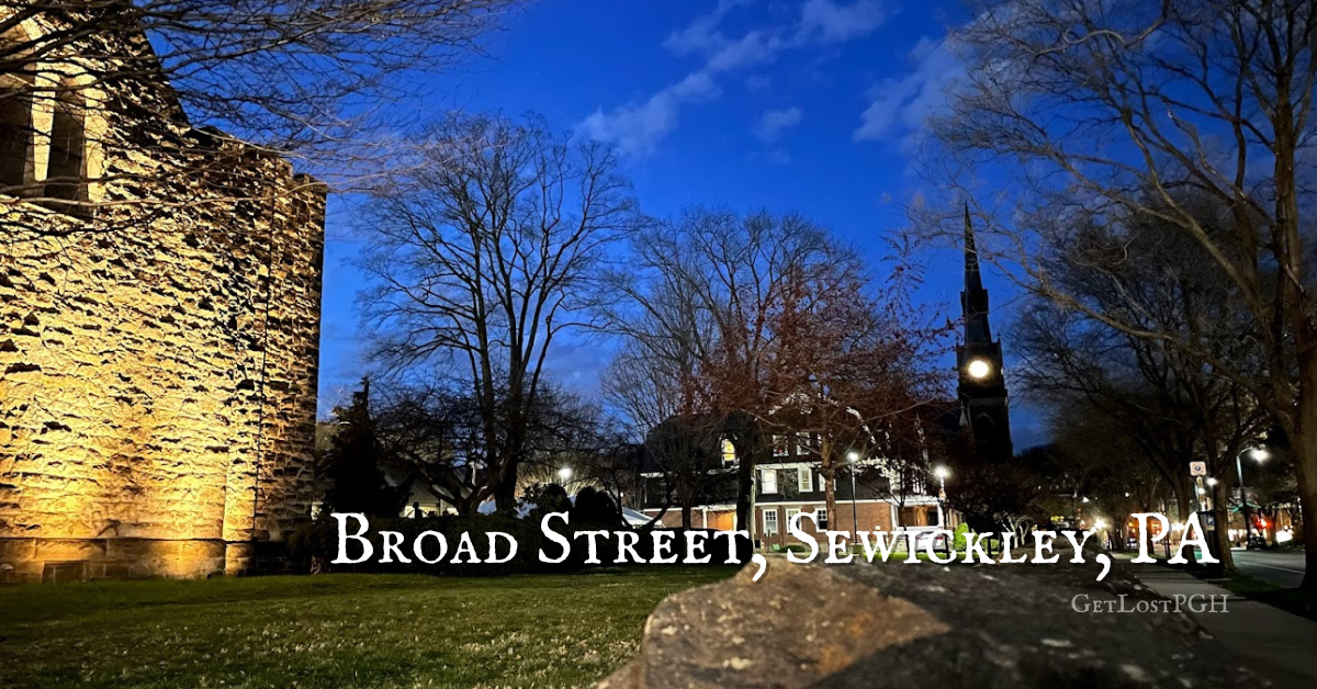 Broad Street in Sewickley, Pennsylvania at dusk, featuring a lit stone church wall, historic homes, and the town clock tower under a deep blue sky. Photo by GetLostPGH.
